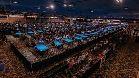 A large indoor venue filled with numerous pool tables where many people are playing or watching a billiards tournament. Description generated by AI